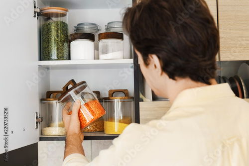 man taking a glass jar of red lentils from a shelf for food storage, organizing grains, rice, and cooking ingredients in pantry jars to prepare healthy meals during home routine.