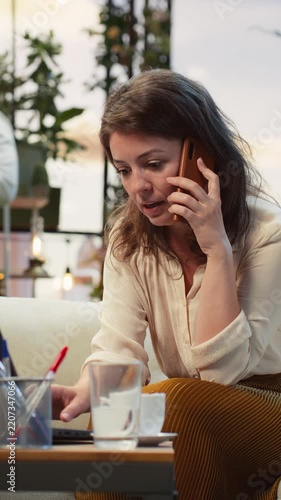 Vertical screen: Businesswoman handles a smartphone call while typing on a laptop keyboard in a rooftop garden, talking on the phone and multitasking with the notebook as a female entrepreneur.
