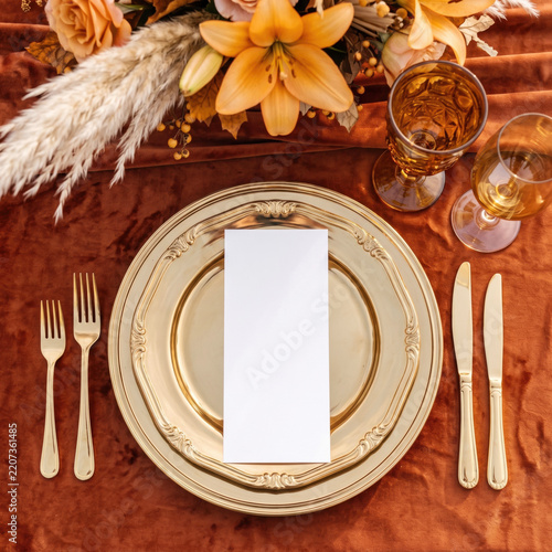 Elegant autumn table setting with a decorative plate, golden cutlery, seasonal flowers and a blank menu mockup arranged on rich rust-colored fabric