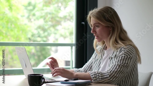 A young blonde woman diligently works on her laptop from home. She takes a thoughtful pause, looking out the window, perhaps pondering an idea or momentarily distracted from her tasks.