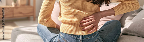 Back Pain Distress: A young person experiences acute lower back pain, clutching their back in discomfort while sitting on a couch at home, highlighting the struggle of chronic pain