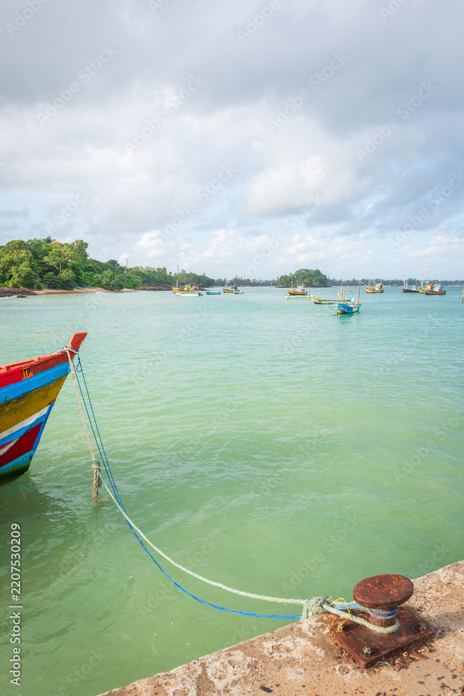Fototapeta premium traditional fishing boat is moored beside a harbor pier in Weligama, Sri Lanka, with ropes secured to a weathered metal bollard in the foreground