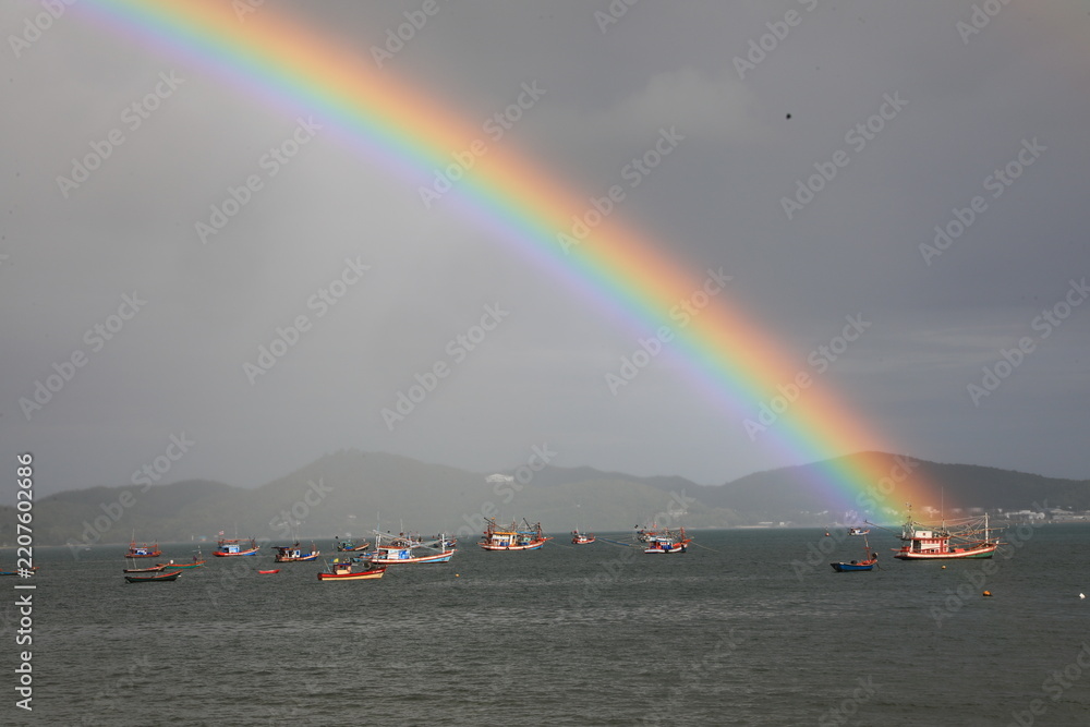Obraz premium A vibrant rainbow arching over the sea with local fishing boats against a dramatic stormy sky in Sattahip, Chonburi, Thailand.