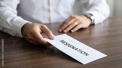 Business person handing a white envelope labeled resignation to a colleague across a wooden office desk with workspace