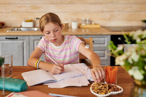 Caucasian kid girl with continuous glucose monitor writing in notebook doing homework and eating healthy snacks. Perfect for diabetes management and pediatric healthcare campaigns