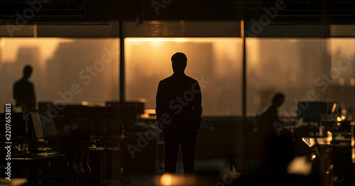 Silhouette in dimly lit corporate office interior during golden hour, creating quiet and reflective workplace atmosphere with blurred workers and warm window light
