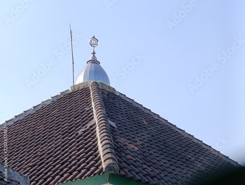 An Islamic symbol finial sits atop a small, silver dome. Brown terracotta tiles form the roof under a clear sky.