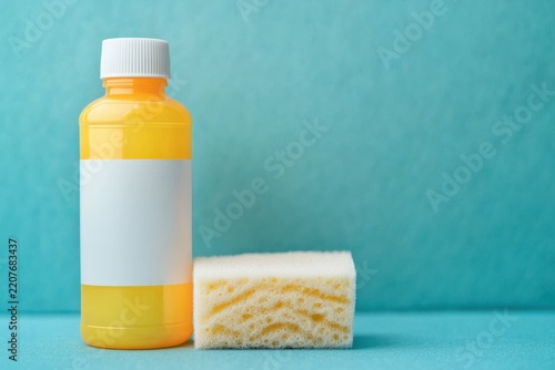 A yellow plastic bottle with a white cap and blank label stands next to a white sponge on a blue surface and background.