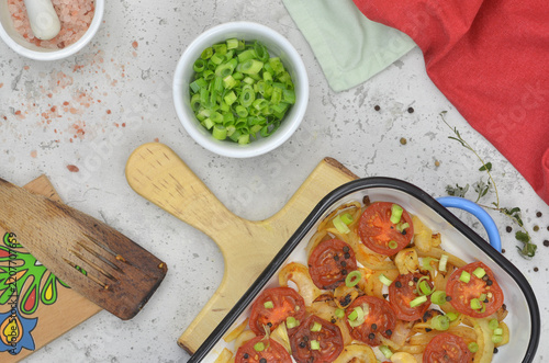 Italian, regional dish with red tomato, onion, garlic and other green herbs. Top view blue roasting pan, chives and salt in bowls, regional pattern on the wooden cutting board.