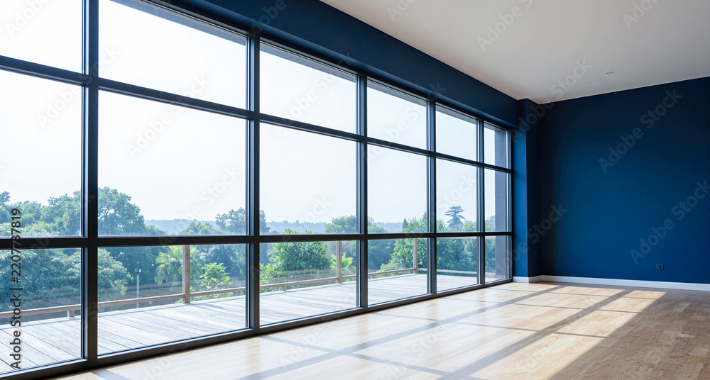 Obraz premium Empty spacious modern room featuring large black framed windows overlooking green trees with dark blue accent wall and wooden parquet floor