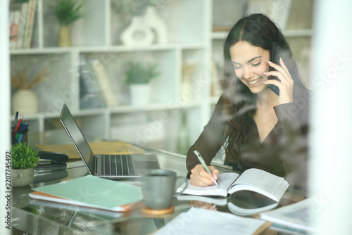 Happy young telecommuter writing in agenda attending phone call