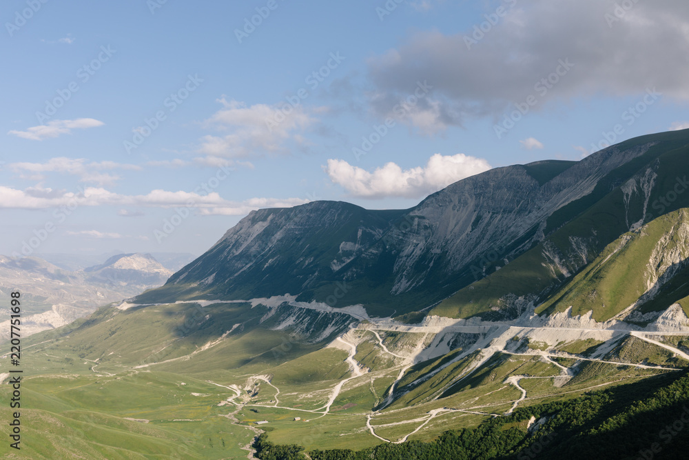 Fototapeta premium Wide panoramic view of the Matlas plateau with rugged mountain ridges, green valleys and winding dirt roads under a blue sky with scattered clouds