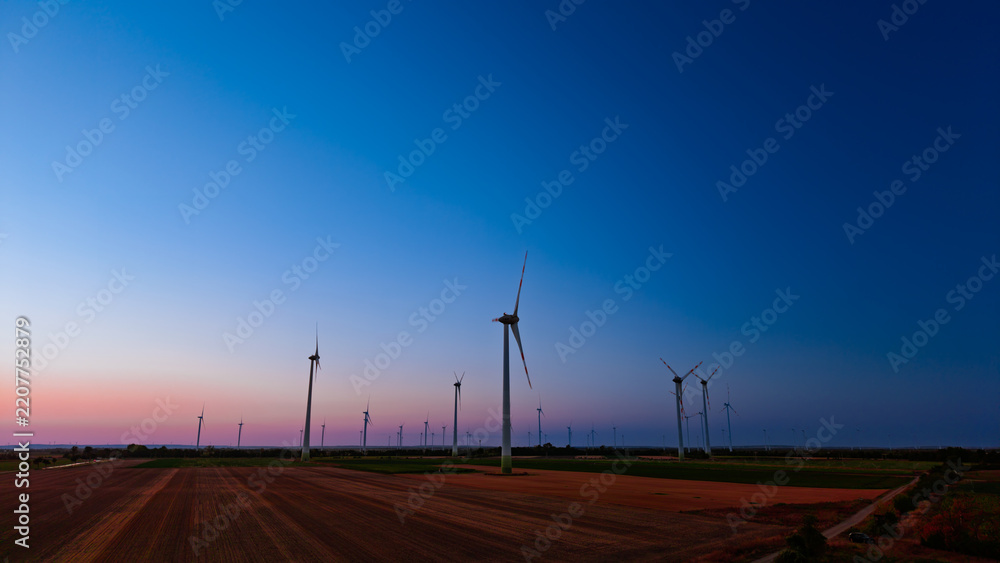 Obraz premium Wind turbines in field at blue hour. Dark generators stand in a wide rural landscape under a clear purple sky at dusk.