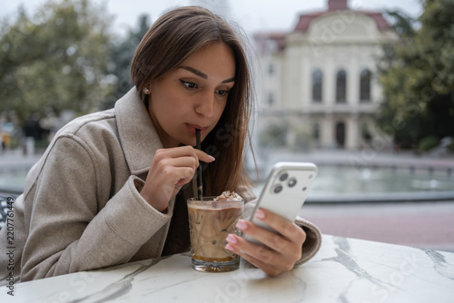 Young woman drinking iced coffee with straw and looking at smartphone at outdoor cafe terrace. Modern lifestyle, authentic emotion, casual tech concept