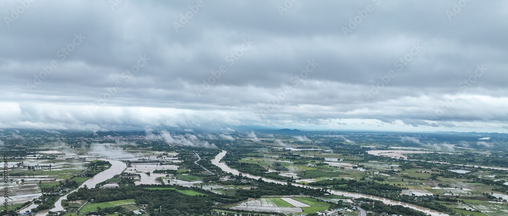 Fototapeta premium Rain clouds blanket swollen river winding through lush agricultural fields, dense mist rolls across the flooded rural landscape in gloomy monsoon day.
