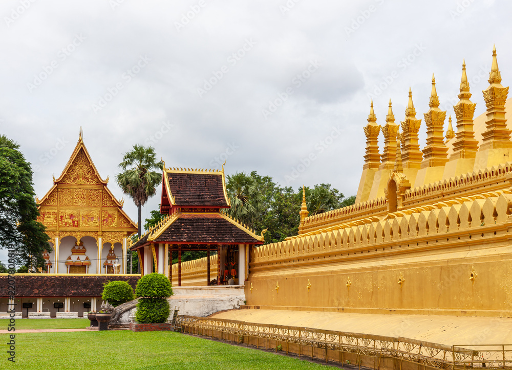 Fototapeta premium Golden stupa and temple complex stand under overcast sky, surrounded by green lawn and paved walkway in Vientiane, Laos.