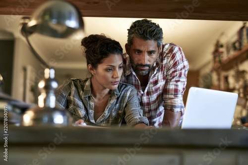 Two adults studying laptop together at home with focused expressions, plaid shirts, warm interior lighting, and blurred foreground, collaborative domestic workspace scene