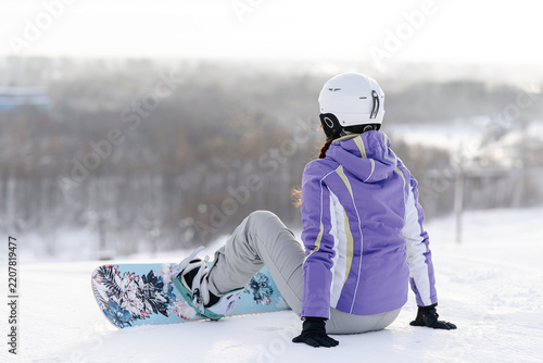 young active caucasian woman snowboarding, purple sports clothes, white helmet, sportive woman sits on snow with white snowboard, cloudy winter day, mountain slope, equipped ski run
