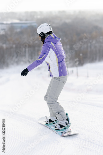 young active caucasian woman snowboarding, purple sports clothes, white helmet, white snowboard, sportive woman stands on snowboard, back view, cloudy winter day, mountain slope, equipped ski run
