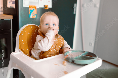 A charming portrait of a sweet infant child learning to self-feed while sitting safely in a white highchair. The baby wears a patterned bib and holds a spoon, with a partitioned silicone suction plate