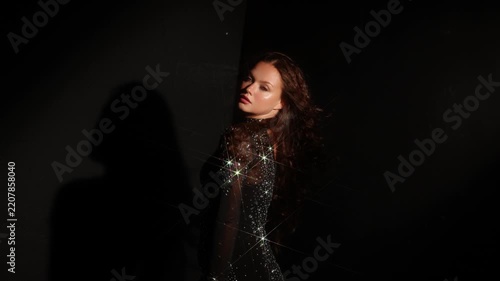 Elegant woman in a shimmering dress with lowered head in the dark. Silver embellishment catches a narrow beam, moody atmosphere of a late party.