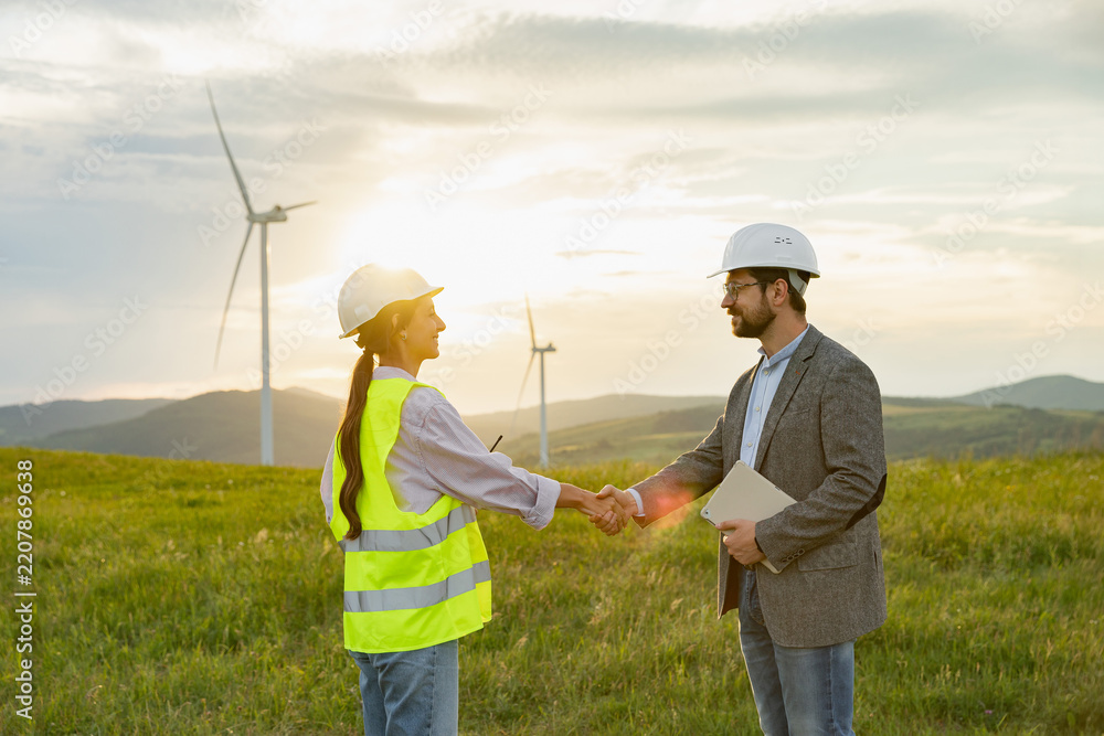 Obraz premium Two engineers in hard hats and safety vests shake hands in a field with wind turbines in the background during sunset