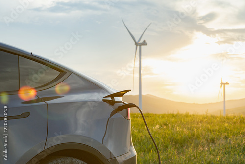 An electric vehicle is plugged into a charging station amidst a field with wind turbines in the background during sunset