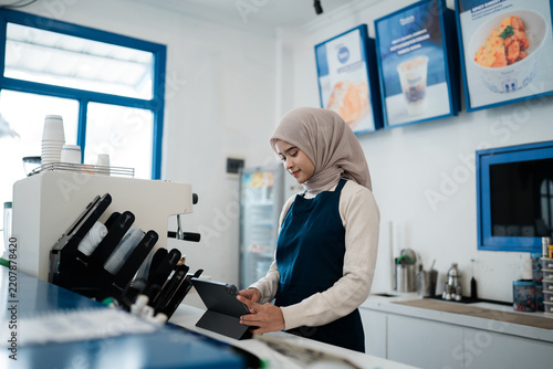 Smiling woman in hijab handles orders on tablet by espresso machine at tidy cafe counter, with menu boards, stacked cups, and blue-framed windows in background.