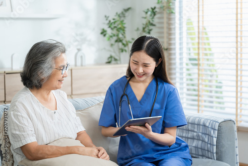 Young nurse in scrubs reviews health records on digital tablet while sitting beside smiling elderly woman on living room sofa during a home health visit.