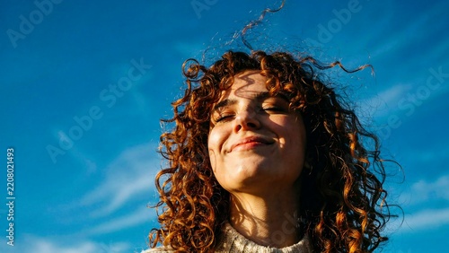 Smiling curly haired woman enjoying sunshine outdoors against a bright blue sky