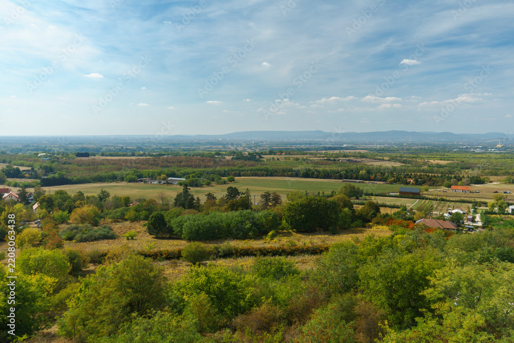 Fototapeta premium Vast panoramic landscape view of rural fields, woodland patches, and distant hills in autumn colors under a bright blue cloudy sky