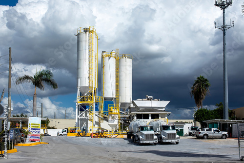 Industrial concrete batching plant factory with tall white storage silos and cement mixer delivery trucks