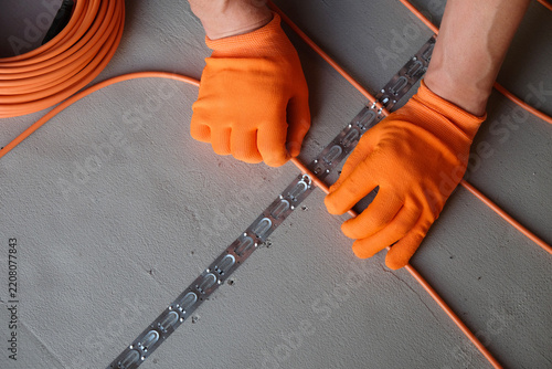 Technician wearing orange gloves attaches a heating cable to the mounting tape on the floor. Close-up view of the underfloor heating installation.