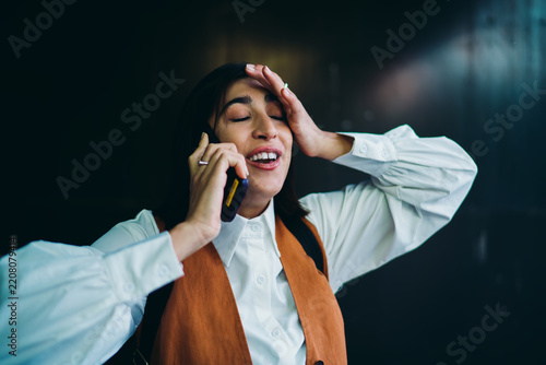 Disappointed latin female student gesturing during cellular conversation network problem nearby dark wall background. Upset hispanic woman holding smartphone touching head with closed eyes.