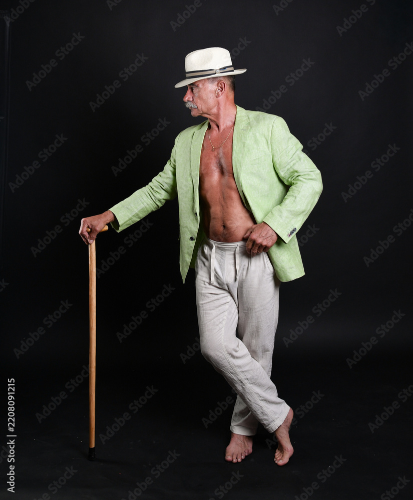 Naklejka premium Exzentrischer älterer Mann mit weißem Hut, grünem Sakko und Spazierstock posiert barfuß vor schwarzem Hintergrund. Charakterporträt, Vintage-Stil, Gentleman, Individualität und kreative Studiofotograf