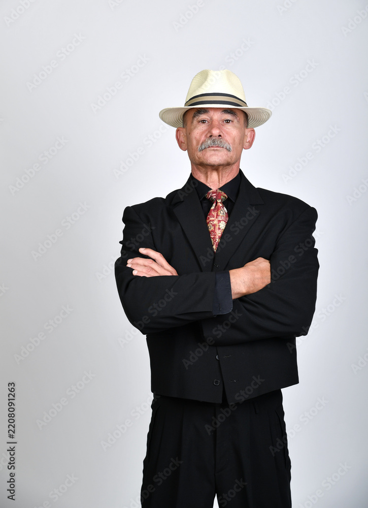 Naklejka premium Selbstbewusster älterer Mann im schwarzen Anzug mit Hut und verschränkten Armen vor neutralem Hintergrund. Charakterporträt, Business, Autorität, Vintage-Stil und elegante Studiofotografie.