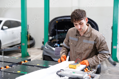 Mechanics working on an electric car in a garage, focusing on battery technology and repair. The foreground shows a mechanic inspecting battery modules with a multimeter.