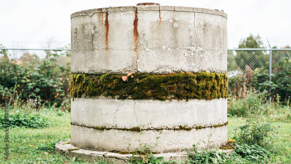 Fototapeta premium Old Concrete Water Tank Covered With Moss And Rust In Outdoor Grass Area