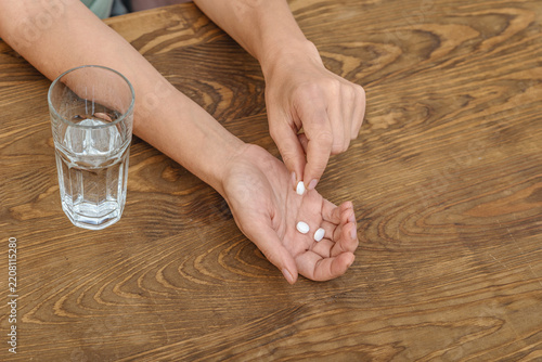 Close-up of woman's hands holding pills and glass of water against wooden surface. Treatment, medicine