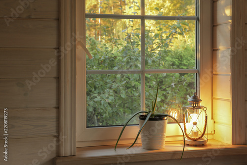 Horizontal cozy window scene with glowing lantern, potted plant, green garden view, warm evening light, wooden interior, brown, green, white tones, peaceful autumn home decor, window background image.