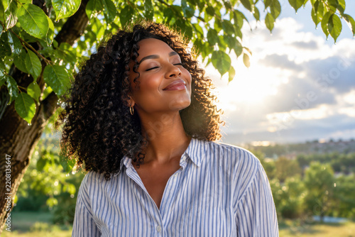 Beautiful young African American woman stands with her eyes closed, smiling peacefully as she enjoys the warm golden hour sunlight filtering through lush green leaves