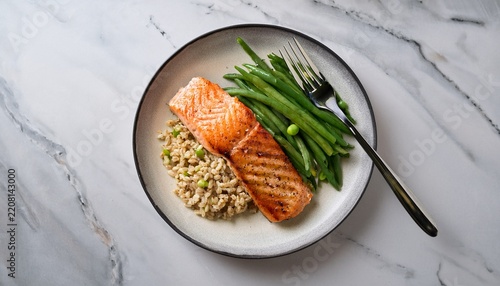 grilled salmon fillet served on a round plate with green beans and barley featuring a fork resting beside the dish on a light marble surface showcasing a healthy meal presentation