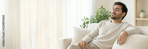 Young man relaxes comfortably on a bright sofa in a modern home, enjoying a calm moment with natural light and plants nearby.