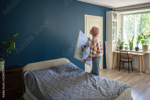 Woman artist holding canvas artwork ready to hang picture over bed on wall in bedroom, creative female painter stands beside bed preparing to place painting in room, wall decor and cozy living space.