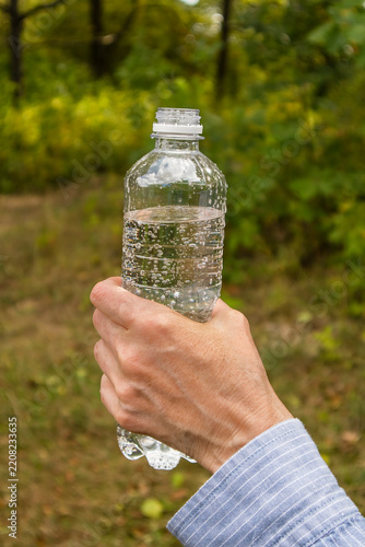 Fotografie Hand holding a reusable-style plastic water bottle on a woodland trail, conveying refreshment, hiking, fitness, and connection with nature