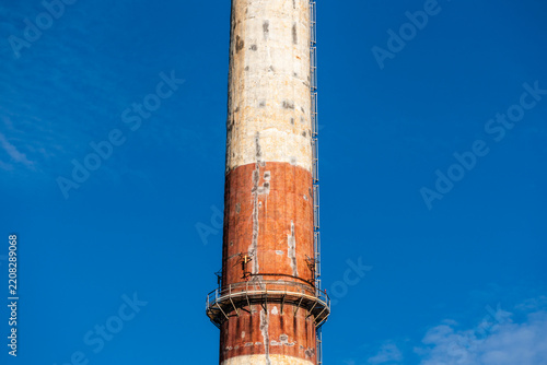 View of a tall industrial chimney with red and white painted concrete, a vertical ladder, and a metal maintenance platform against a clear blue sky in Poland.