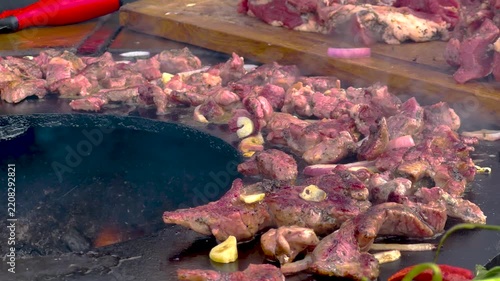 Sizzling pieces of seasoned meat grilling on a hot circular outdoor barbecue plate with rising smoke and sliced red onions during a culinary cookout feast. 4K video.