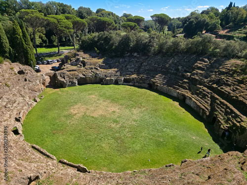 High angle view of the ancient Roman amphitheater of Sutri entirely carved into a tufa rock cliff with green grass arena under a sunny sky, Viterbo, Lazio, Italy