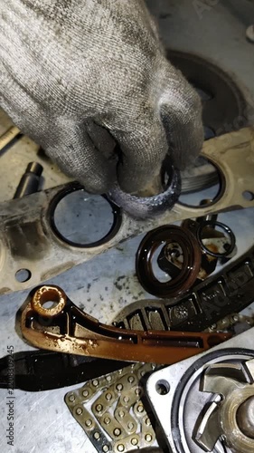 A mechanic in a dirty work glove holds a badly worn, frayed engine seal over a workbench filled with disassembled car parts.