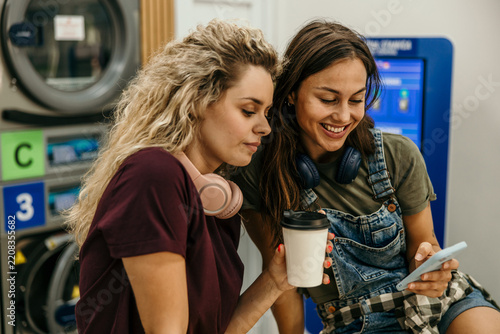 Female friends enjoying coffee and smartphone at laundromat